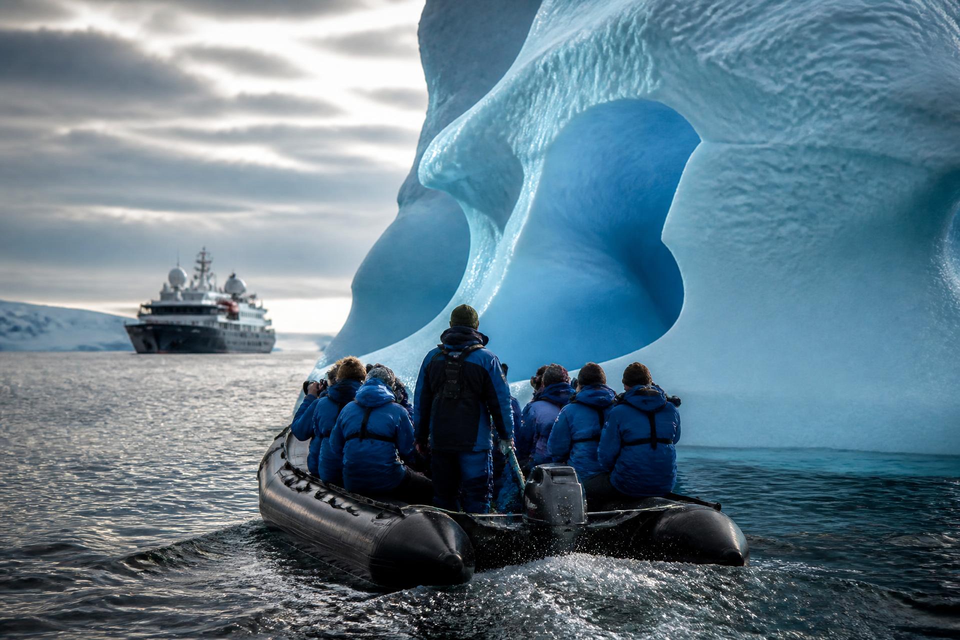 Expedition guests in parkas ride a Zodiac inflatable past a sculpted blue iceberg with their small expedition ship in the distance