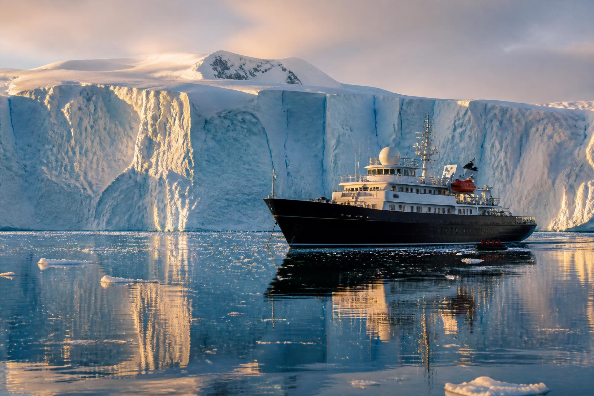An HX Expeditions polar ship anchored in mirror-calm water beside a towering blue glacier wall at golden hour