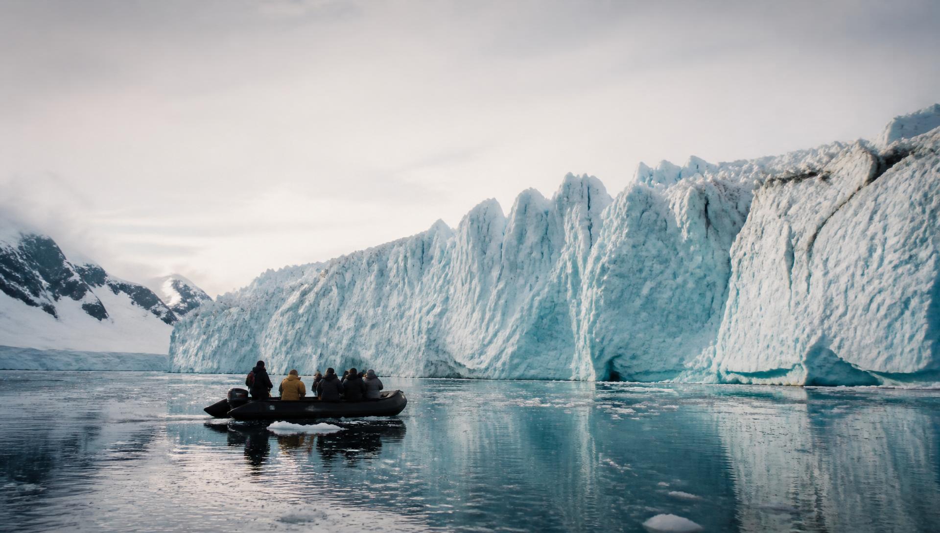 Lindblad-style expedition ship anchored in a remote icy bay at golden hour, with a zodiac of explorers in red parkas approaching shore
